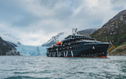 Magellan Explorer dark blue small ship sits in calm water by blue glacier on cloudy day in Patagonia.
