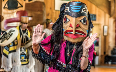 Woman in pink robe with black fringe & native carved wooden mask dances for a cultural presentation on the Remarkable Journey.