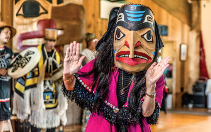 Woman in pink robe with black fringe & native carved wooden mask dances for a cultural presentation on the Remarkable Journey.
