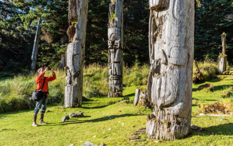 Man in red jacket stands in grass & photographs weathered totem poles on an Alaska, BC & Canada small ship cruise.