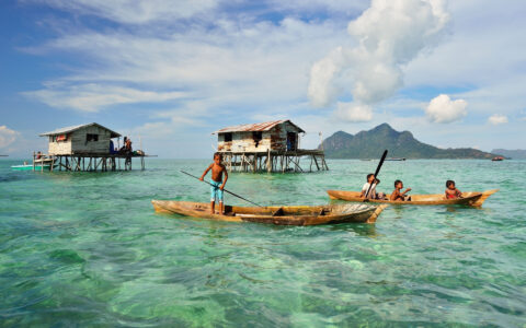 Kids in sea gypsy village Bajau Laut, Indonesia, on a wooden boat in front of houses on stilts over emerald water on a sunny day.