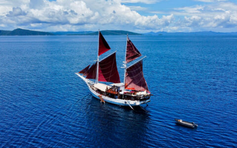Aerial view of small white sailing ship with 5 burgundy sails open & small dinghy towed behind, in calm blue Indonesian sea.