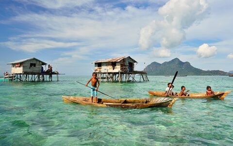 Kids in sea gypsy village Bajau Laut, Indonesia, on a wooden boat in front of houses on stilts over emerald water on a sunny day.
