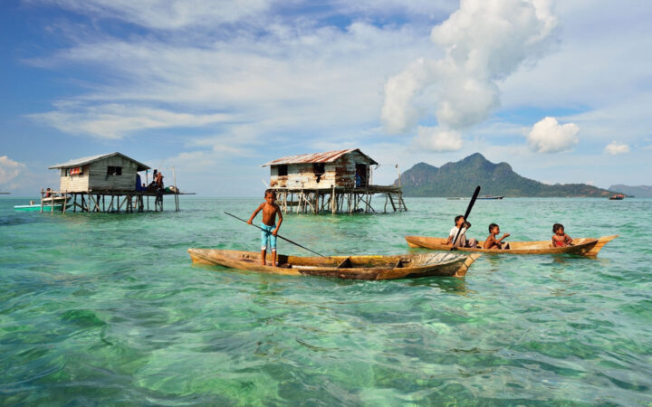 Kids in sea gypsy village Bajau Laut, Indonesia, on a wooden boat in front of houses on stilts over emerald water on a sunny day.