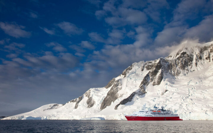 Small polar expedition ship with red hull & white upper decks sits in glassy sea beside tall, snowy cliffs in Antarctica.