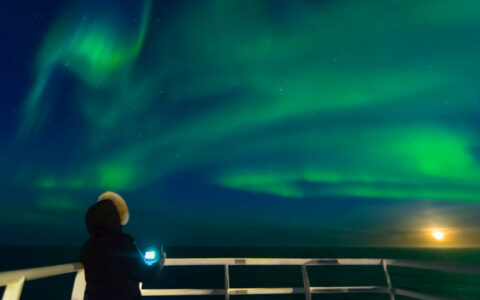 Arctic traveler stands on deck of a small ship at night, photographing wavy green northern lights as the sun sets on the horizon.