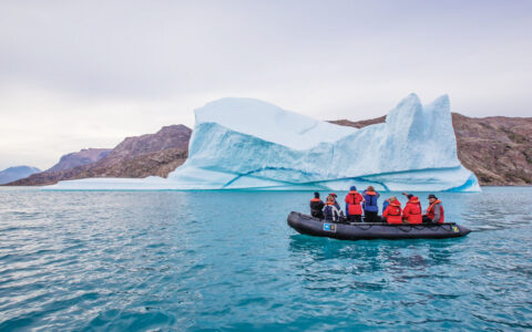 Black inflatable Zodiac dinghy with Arctic travelers motors past a large iceberg in calm water on an Iceland to Greenland cruise,