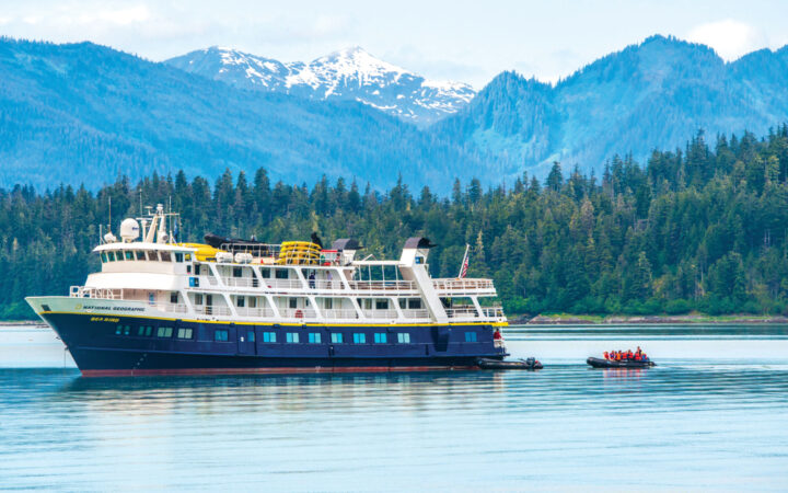 Nat Geo Sea Bird cruises in front of a forested hillside and rocky mountain range, followed by guests in an inflatable skiff.