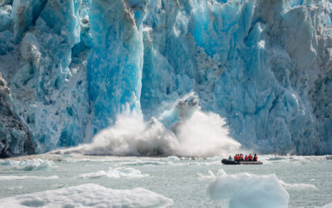Zodiac dinghy with Wild Alaska Escape cruise travelers motors past an icy blue glacier as it calves & creates a large splash.