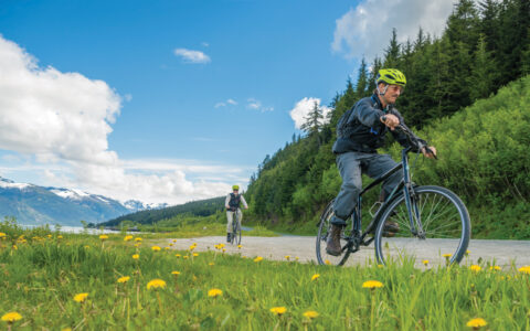 2 men on the Wild Alaska Escape cruise ride black bikes on pavement & dirt along grassy fields beside a river in Haines.