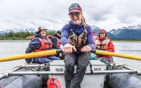 Young woman smiles while rowing a boat full of Wild Alaska Escape travelers down the Chilkat River with mountains behind.
