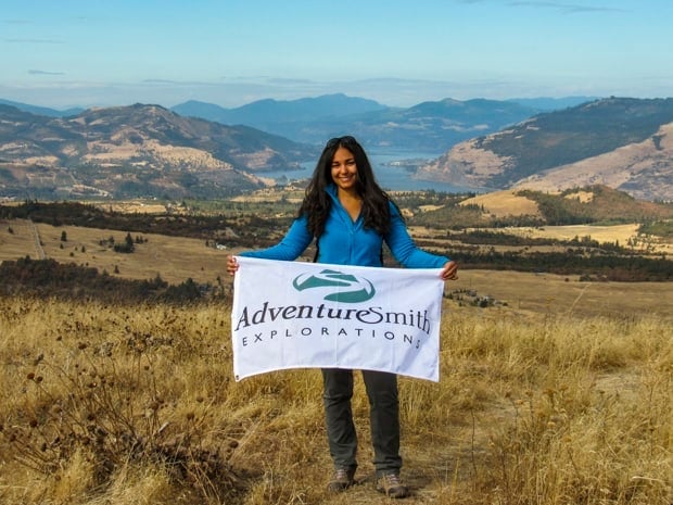A woman with dark hair and a blue shirt holds an AdventureSmith Explorations white flag while standing above the Columbia River