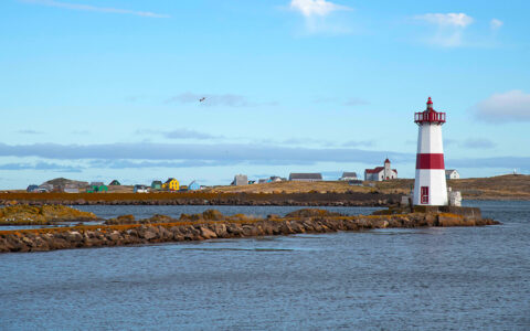 Red & white lighthouse sits on rocky sea wall by calm ocean under blue skies seen on a St. Lawrence river cruise.