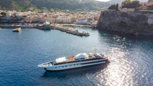 Aerial view of Harmony G white & black mega yacht with 3 guest decks sitting at anchor in Mediterranean port on a sunny day.