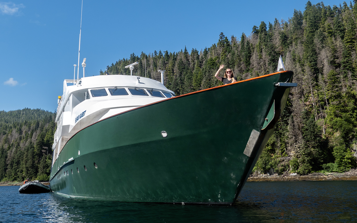 A solo blonde female traveler waves from the bow of a small cruise ship in Alaska