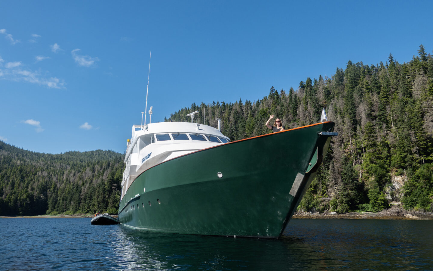A solo blonde female traveler waves from the bow of a small cruise ship in Alaska