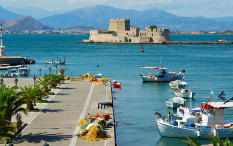 Turquoise sea with a beige castle in the background & paved promenade along the water in the foreground, in Nafplion, Greece.