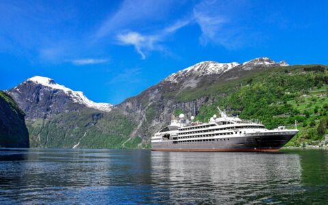 Small expedition ship with gray hull & white upper decks cruises in glassy water by lush green mountains with snowy peaks.