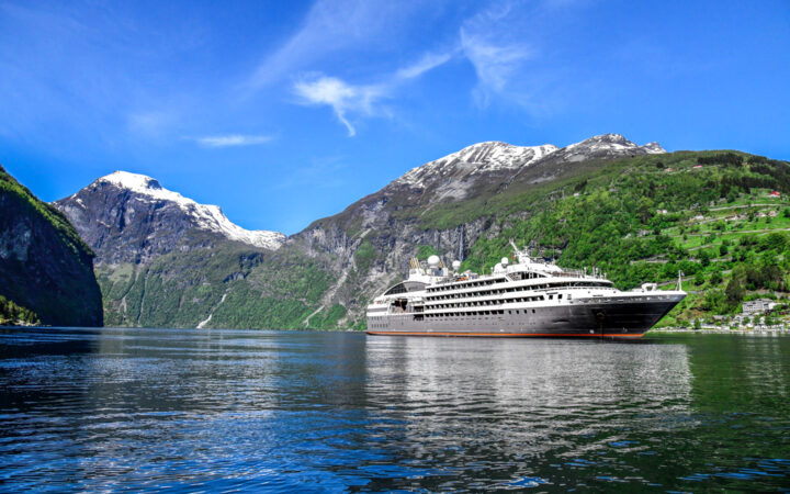 Small expedition ship with gray hull & white upper decks cruises in glassy water by lush green mountains with snowy peaks.