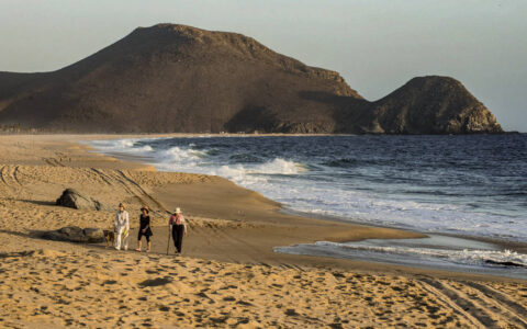 three people walking along the ocean on a sandy beach as the sun is starting to go down