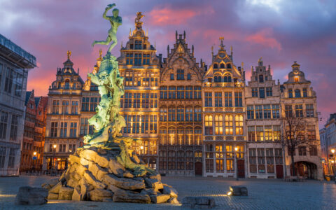 Green statue lit up in front of glowing gothic buildings under a pink & purple sky, seen on a Belgium river cruise.