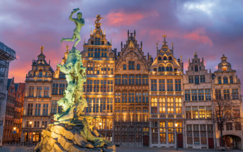 Green statue lit up in front of glowing gothic buildings under a pink & purple sky, seen on a Belgium river cruise.