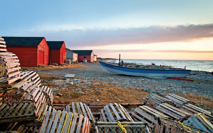 A blue wooden boat, bright red houses and wooden crab pots set along the rocky shoreline of Newfoundland during a pastel sunset.