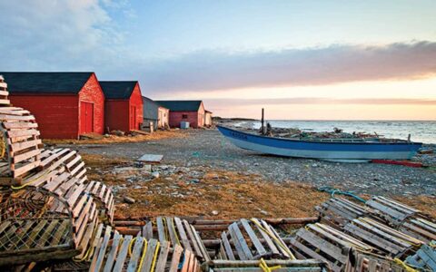 A blue wooden boat, bright red houses and wooden crab pots set along the rocky shoreline of Newfoundland during a pastel sunset.
