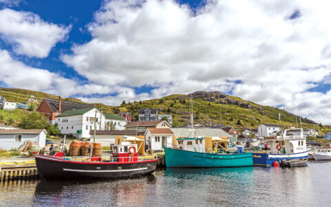 Colorful wooden fishing boats sit tied along shoreline of small village backed by green hills in the Canadian Maritimes.