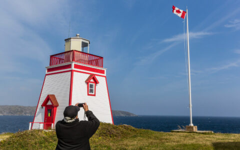 Man in black jacket photographs Canadian flag flying beside red & blue lighthouse on a Canadian Maritimes cruise.