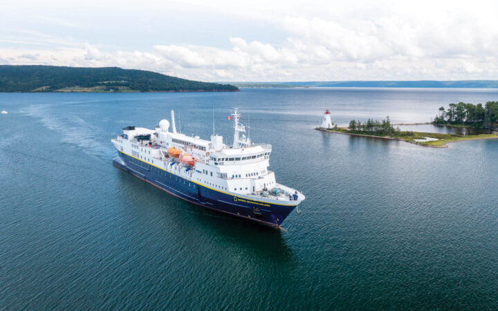 Blue & white Nat Geo ship cruises past small red & white lighthouse in an inlet among green hills in Canada.