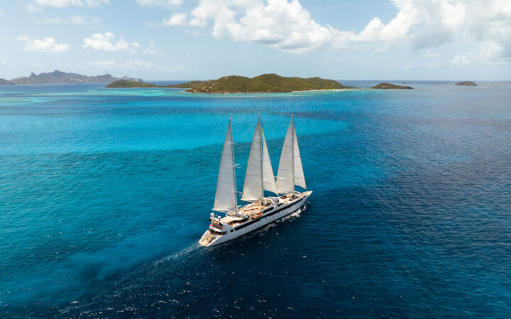 Aerial view of Le Ponant white sailing ship with 3 masts cruising through turquoise sea on a sunny day in the Caribbean.