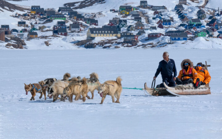 Dog sled with Inuit man & 2 Greenland travelers in orange parkas is pulled across snow by a team of dogs with thick white fur.