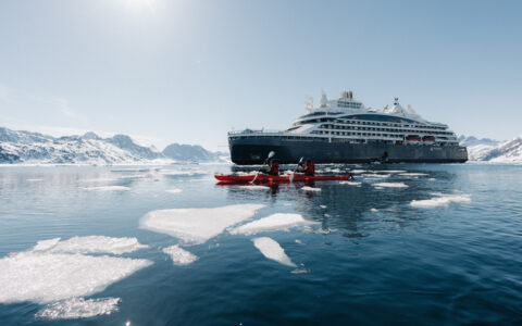 Le Commandant Charcot small ship with blue hull & white upper decks sits in sunshine while tandem kayakers paddle red boats by ice.