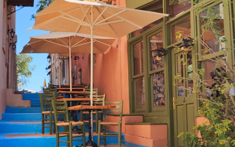 Al fresco cafe dining tables set up under beige umbrellas atop blue steps in Greece.