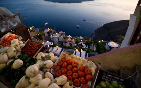Vegetables laid out in boxes look down on travelers dining on a terrace overlooking calm seas at sunset in Greece.