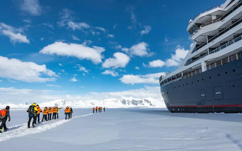 Le Commandant Charcot is parked in an ice sheet in Bellingshausen sea, travelers walk through snow on the ice around it,