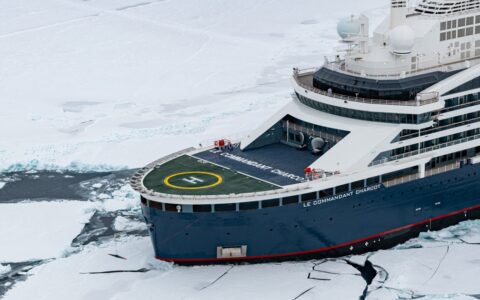 Aerial view of modern expedition ship with dark blue hull, white upper decks & helicopter pad on its bow, parked in ice at the North Pole.