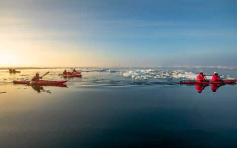 Tandem kayakers in red boats paddle glassy water among small icebergs at dusk on an Arctic cruise near the North Pole.