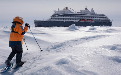 North Pole traveler in orange jacket snowshoes in a windswept area with Le Commandant Charcot polar ship in the distance.