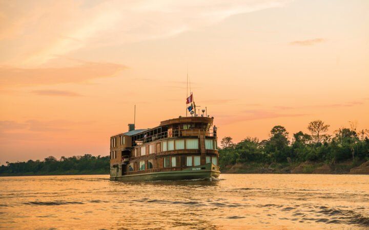 Delfin II Amazon river boat cruising down the calm river enveloped in an orange and pink sunset backdrop. Its 3 decks stand tall on the water.