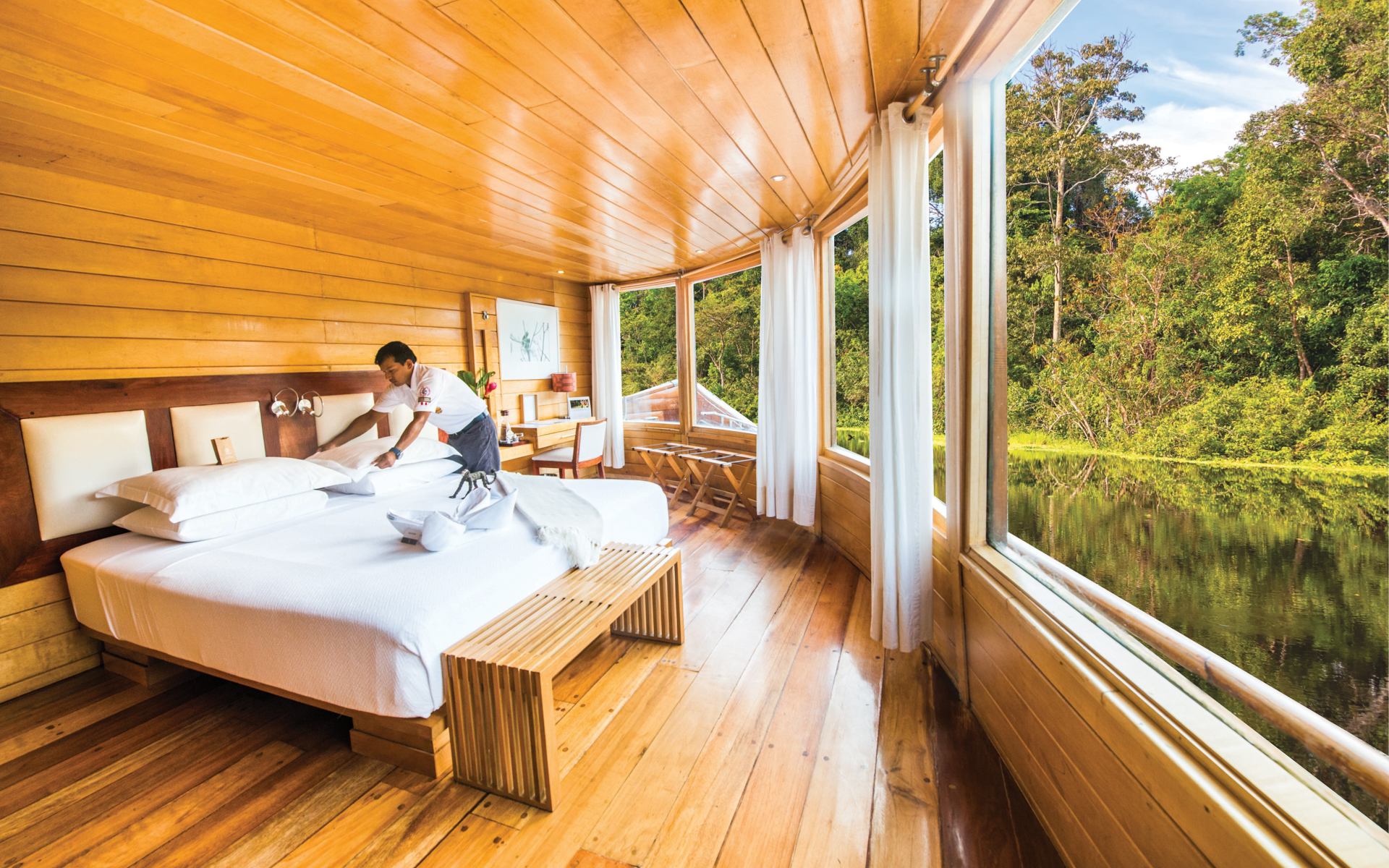 Cabin steward makes up king bed by wraparound picture windows in Amazon Suite of Delfin II riverboat on an Amazon River cruise.