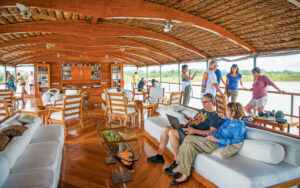 Guests sit on couch & stand along open-air railings in al fresco lounge with couches, chairs & tables on Delfin II riverboat.