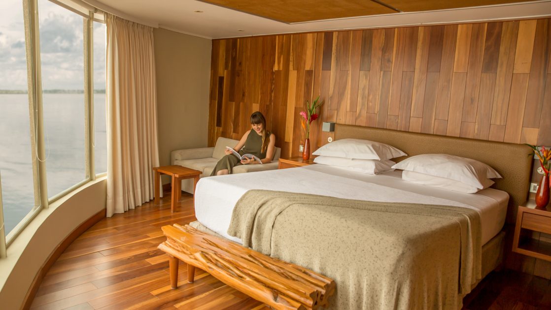Corner Suite of Delfin III Amazon riverboat with wraparound floor-to-ceiling windows, wood finishes & woman on couch beside double bed.