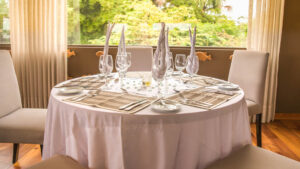 Round 4-top table set with white linens, beige placements & wine glasses with tan napkins beside window on Delfin III ship.