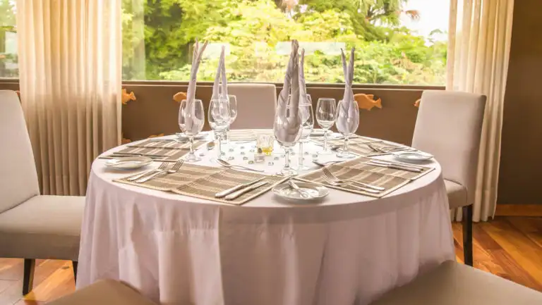 Round 4-top table set with white linens, beige placements & wine glasses with tan napkins beside window on Delfin III ship.
