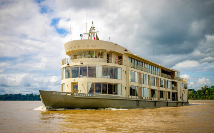 Exterior of bow & port side of Delfin III riverboat with green hull & beige upper decks cruising in the Peruvian Amazon.
