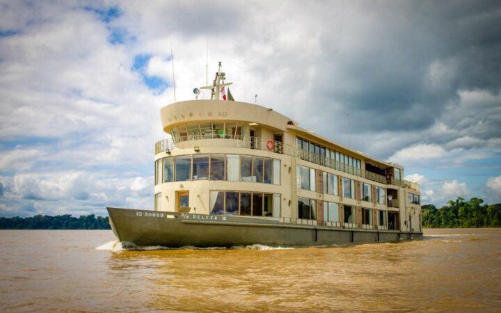 Exterior of bow & port side of Delfin III riverboat with green hull & beige upper decks cruising in the Peruvian Amazon.