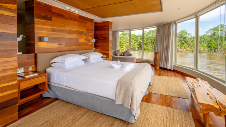 Owner's Suite on Delfin III Amazon riverboat with wood finishes, wraparound windows & large double bed in white linens.