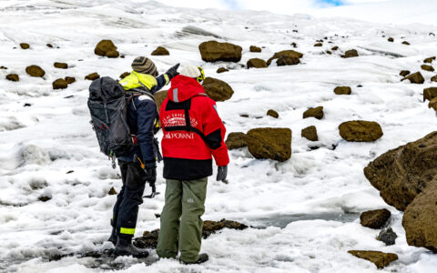 Guide with backpack & Antarctica traveler in red jacket stand atop snow & boulders field, looking around on a cloudy day.